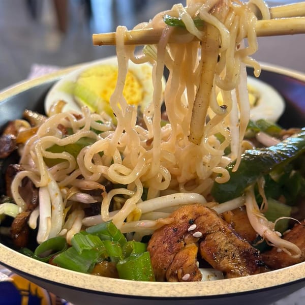 Chopsticks lifting ramen noodles from a bowl with chicken, bean sprouts, green onions, and egg.