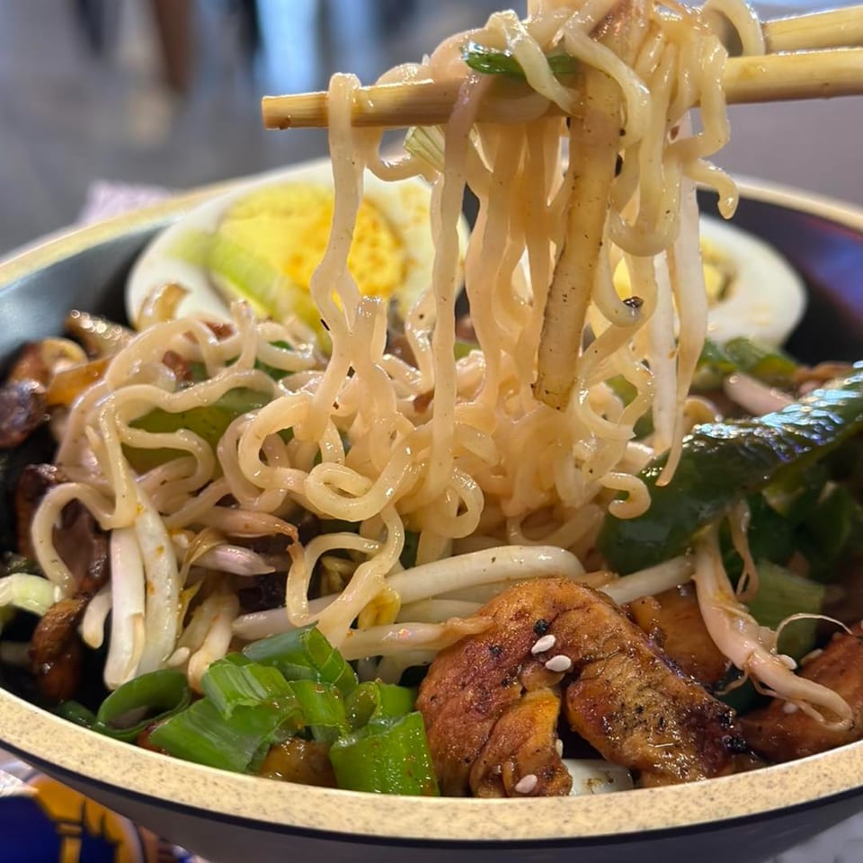 Chopsticks lifting ramen noodles from a bowl with chicken, bean sprouts, green onions, and egg.